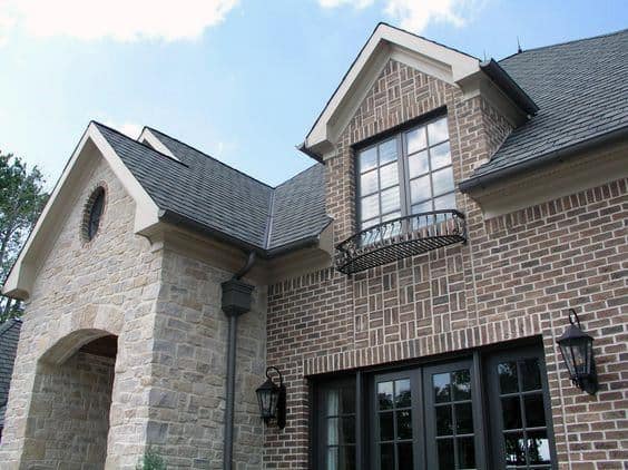 Elegant home featuring intricate brick and stone patterns, wrought iron balcony, black-framed windows, and classic lantern sconces