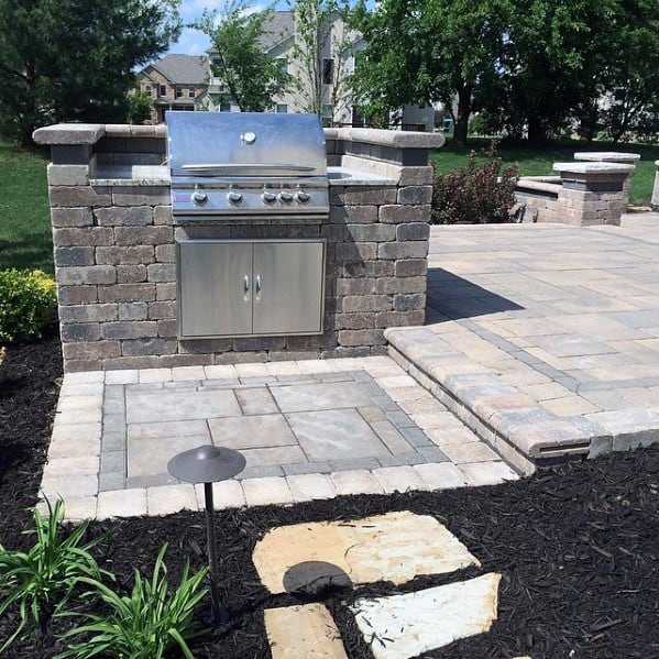 Built-in brick grilling station with stainless steel grill on a paved patio, surrounded by lush greenery