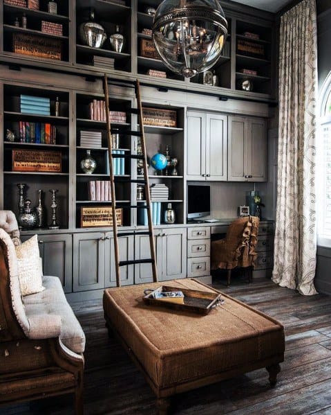 Cozy home library with gray bookshelves, a ladder, brown ottoman, and desk by a large window