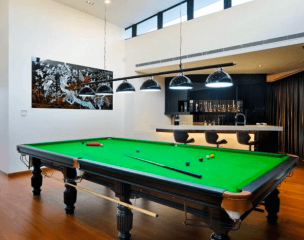 Modern billiard room with green felt pool table, bar area, and large black pendant lights.