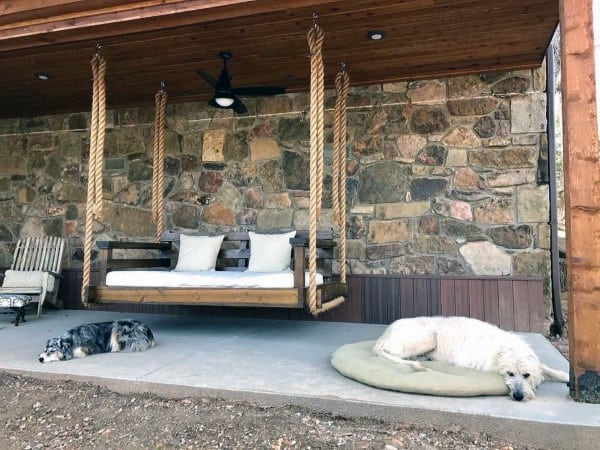 Rustic hanging bench with thick rope supports, stone wall backdrop, and relaxed outdoor setting.
