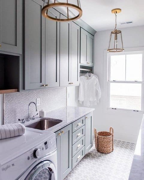 Modern laundry room with gray cabinets, brass accents, patterned flooring, and natural light.