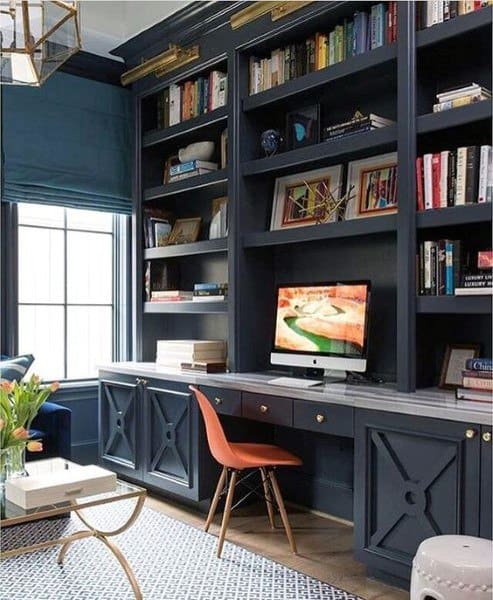Home office with navy blue built-in shelves, a wooden desk, and an orange chair, featuring a computer and books