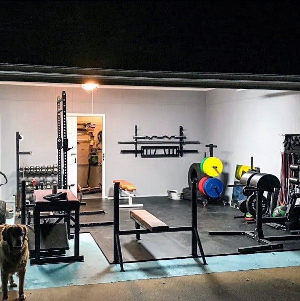 Garage gym with squat rack, bench press setup, weight plates, dumbbells, and GHD machine on rubber flooring.