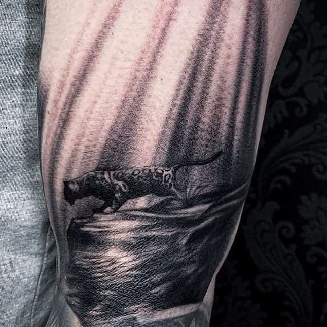 A realistic black and grey tattoo of a jaguar walking on a rock, with soft shading and dramatic light filtering from above, placed on the forearm.