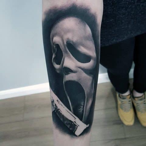 Realistic black and grey tattoo of the Ghostface mask from *Scream*, featuring dramatic shading and a bloodied knife for a chilling horror effect.