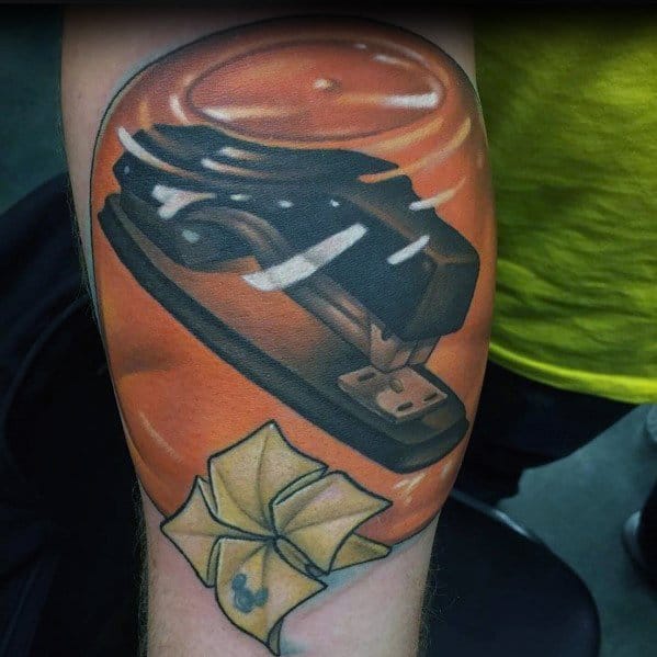 Color tattoo of a black stapler encased in orange Jell-O, referencing Jim's classic prank on Dwight from The Office, with a folded note below.