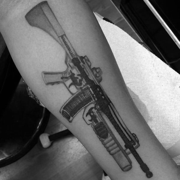 Black and grey AR-15 tattoo on the forearm, featuring detailed shading, a tactical grip, and a grenade launcher attachment, symbolizing strength and precision.