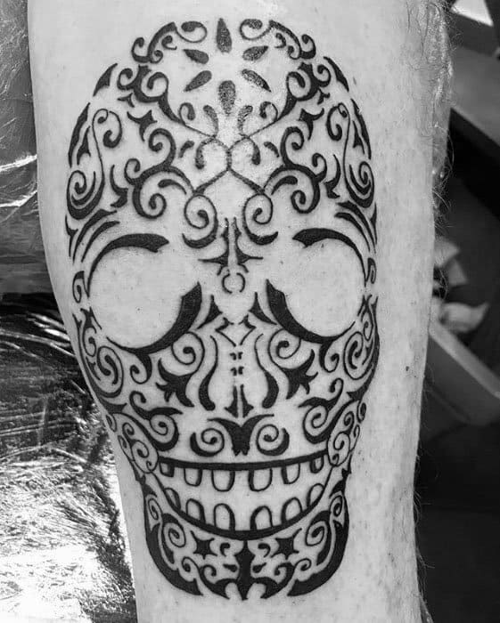 Tribal sugar skull tattoo on leg, featuring ornate swirls and symmetrical patterns in bold black ink for a decorative, festive look.