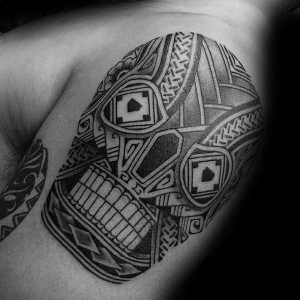 Polynesian-style tribal skull tattoo on upper arm, detailed with geometric patterns, bold lines, and symmetrical tribal motifs.