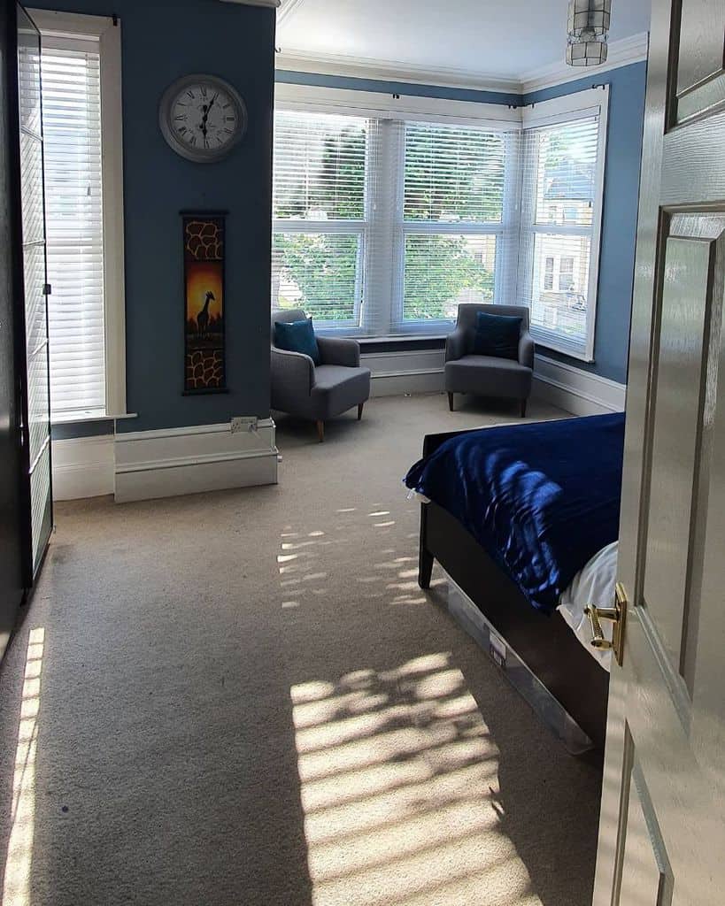 Spacious blue bedroom with large windows, grey chairs, and a navy bedspread.