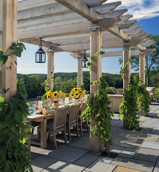 Light wood pergola with climbing greenery, lantern lights, and a long dining table set with sunflowers.