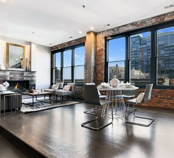 Open-concept industrial apartment with brick walls, large windows, and modern dining furniture.