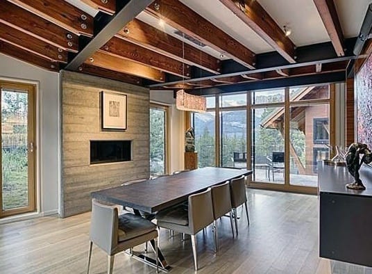 Modern dining room with a concrete fireplace, wooden beams, large windows, and a sleek table.