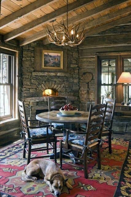 Cozy log cabin dining room with stone corner fireplace, antler chandelier, and colorful rug.