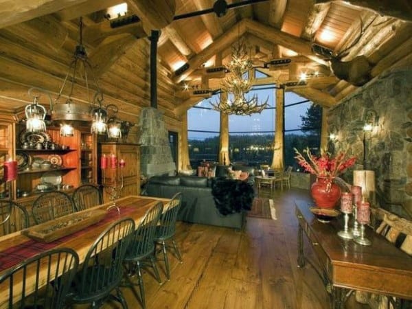 Rustic log cabin dining area with vaulted ceilings, antler chandelier, and cozy stone fireplace.