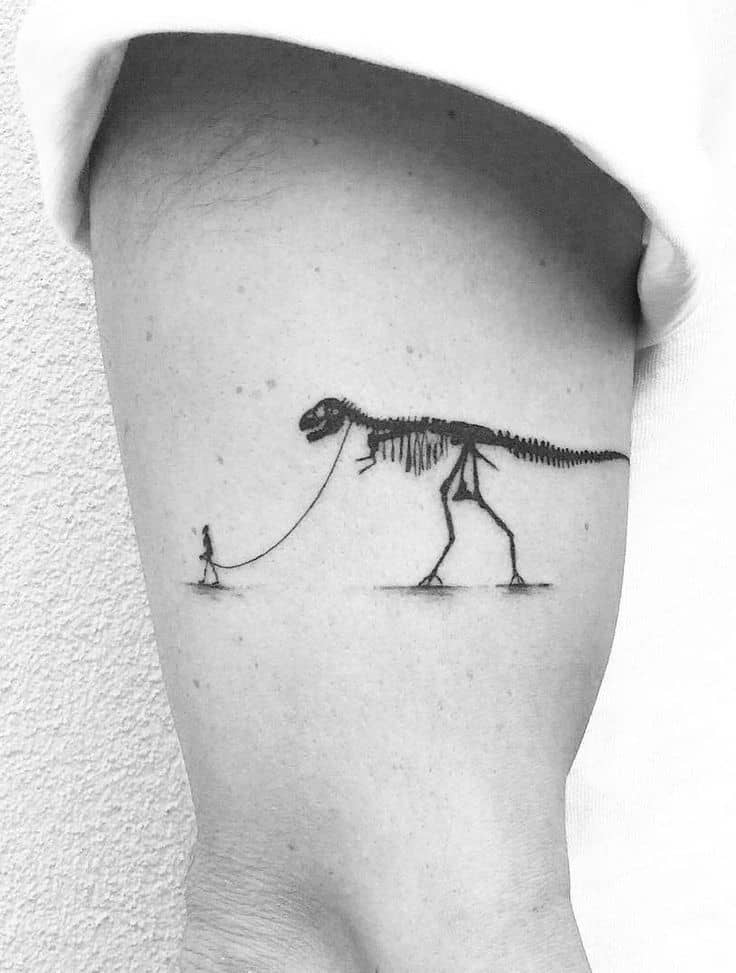 A black ink tattoo of a tiny person walking a massive T-Rex skeleton on a leash, blending surrealism with a minimalist design on the arm.