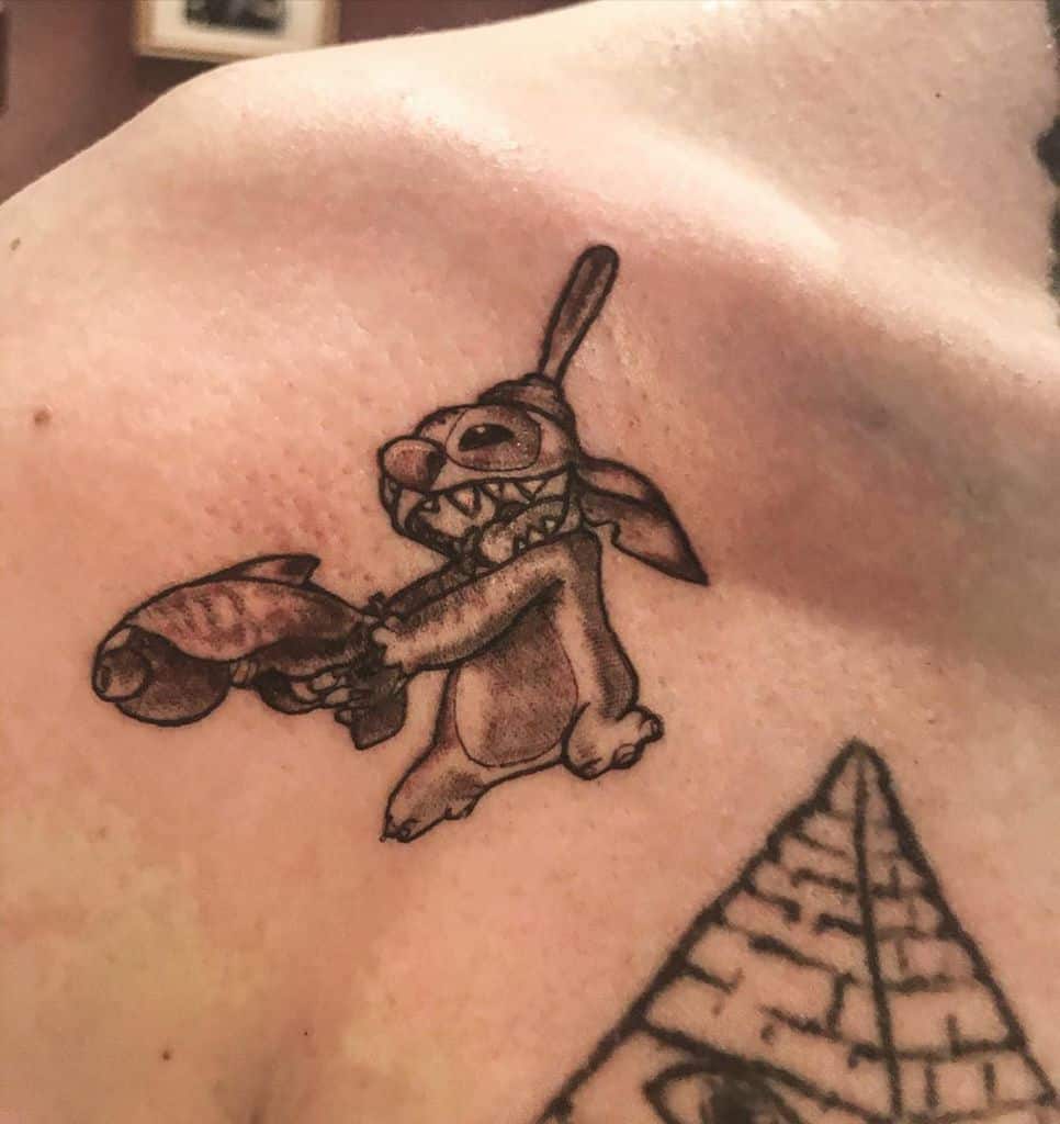 Black and gray tattoo of Stitch from Lilo & Stitch holding a fish with a mischievous grin, located on the shoulder.
