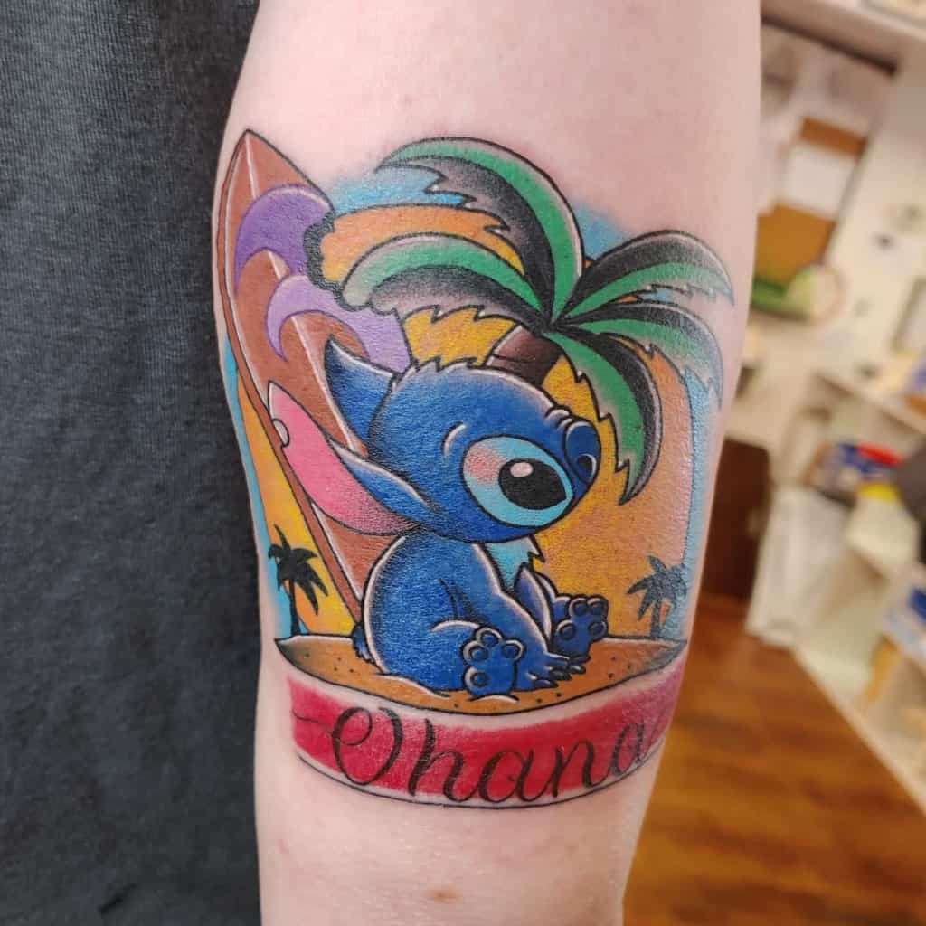 Colorful tattoo of Stitch from Lilo & Stitch sitting by a palm tree with a surfboard, and the word 'Ohana' below, on the forearm.