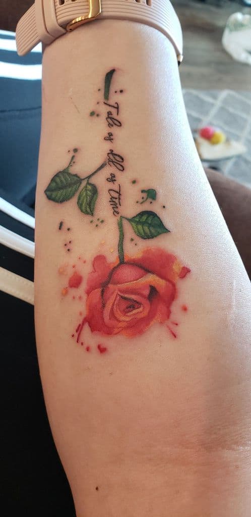 Watercolor-style tattoo of a falling enchanted rose with 'Tale as Old as Time' written along the stem, blending soft reds and greens.