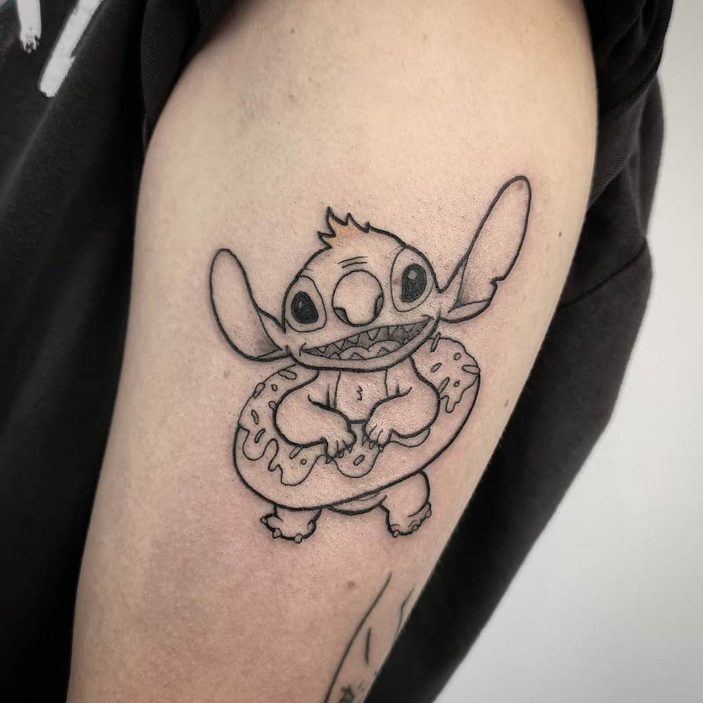 Black and white tattoo of Stitch from Lilo & Stitch holding a donut with icing, placed on the upper arm.