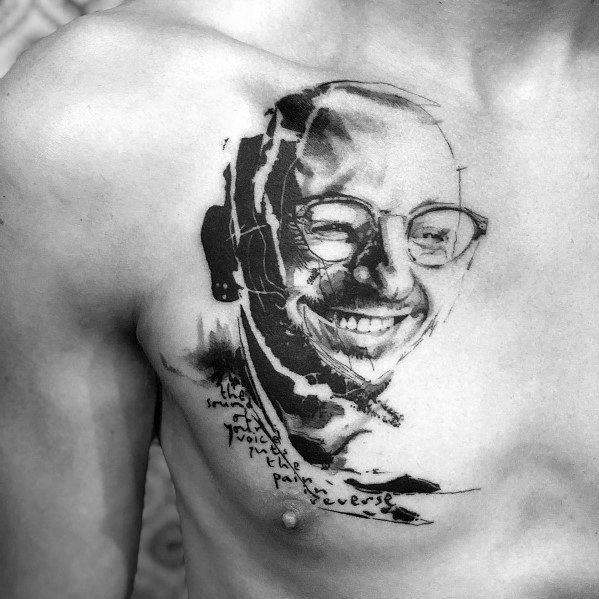 Artistic black ink chest tattoo of Chester Bennington, featuring a smiling portrait with abstract brush strokes and handwritten lyrics.