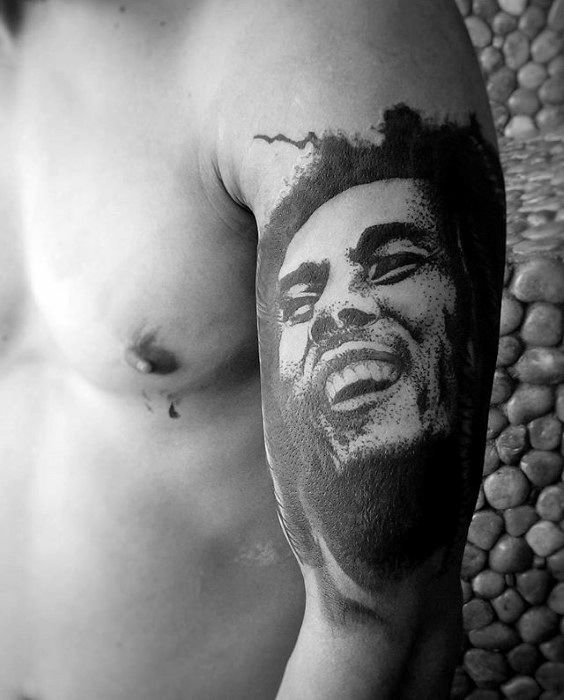 Upper arm tattoo of Bob Marley smiling, done in a bold black ink style with soft shading and textured contrast for a striking portrait look.