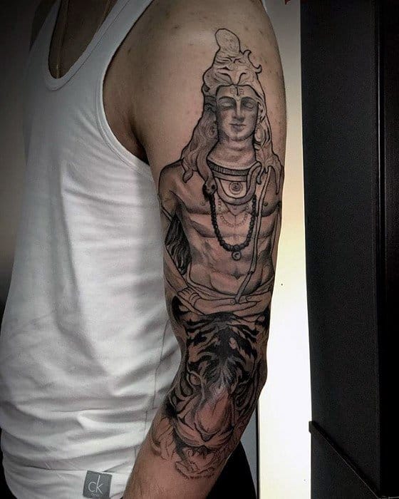 Upper arm tattoo of Lord Shiva with a calm expression, detailed ornaments, and flowing hair, blending into a fierce tiger face on the forearm.