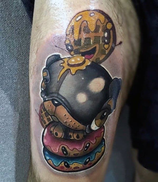 Tattoo of a cartoon-style bear wearing a helmet, stacked on colorful donuts with dripping syrup and a smiling waffle in the background.