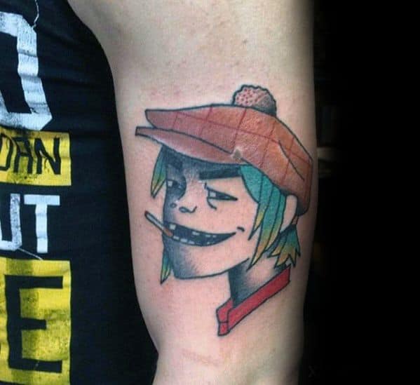 Color tattoo of Murdoc from Gorillaz on the upper arm, with green hair, a smirk, cigarette, and a red beret-style hat.
