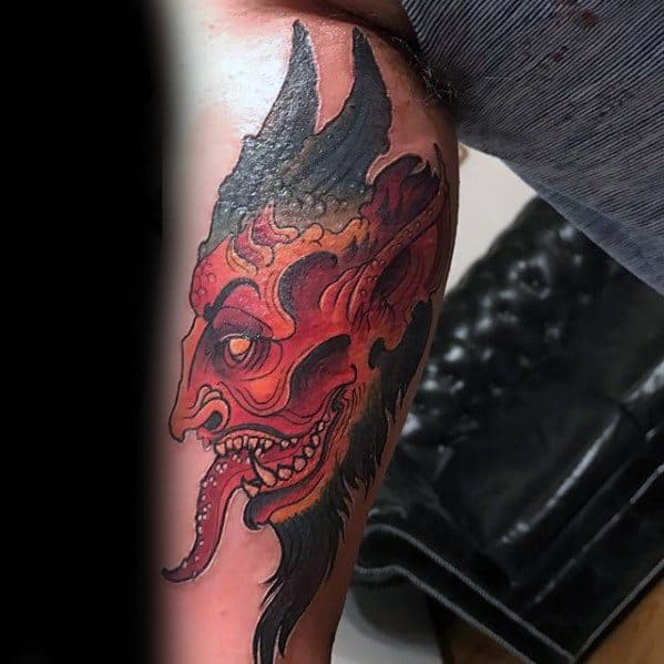 Fiery red and black Krampus tattoo with sharp teeth, pointed ears, and a long tongue, blending demonic and traditional tattoo styles.