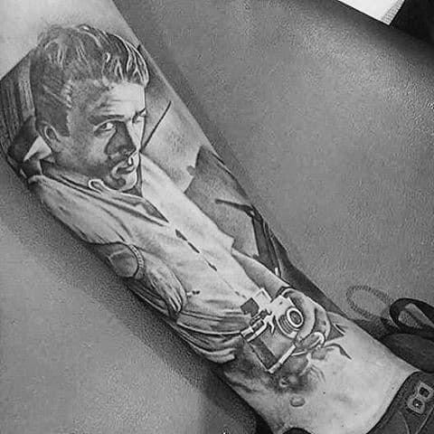 Detailed black and grey tattoo of James Dean holding a vintage camera, with a confident look and retro outfit.