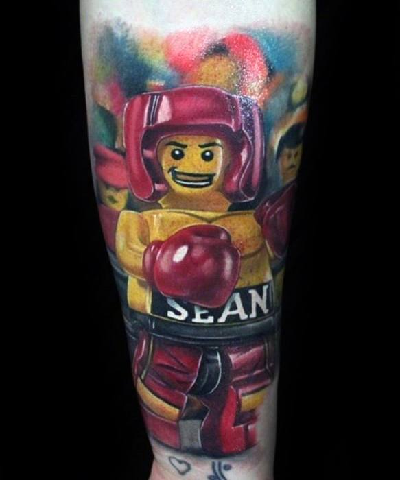 Colorful forearm tattoo of a Lego boxer named Sean, wearing red gloves and headgear, set against a blurred cheering crowd background.
