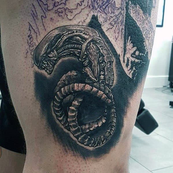 Black and grey Xenomorph tattoo featuring the creature curled in a fetal position with dark shading and detailed textures.