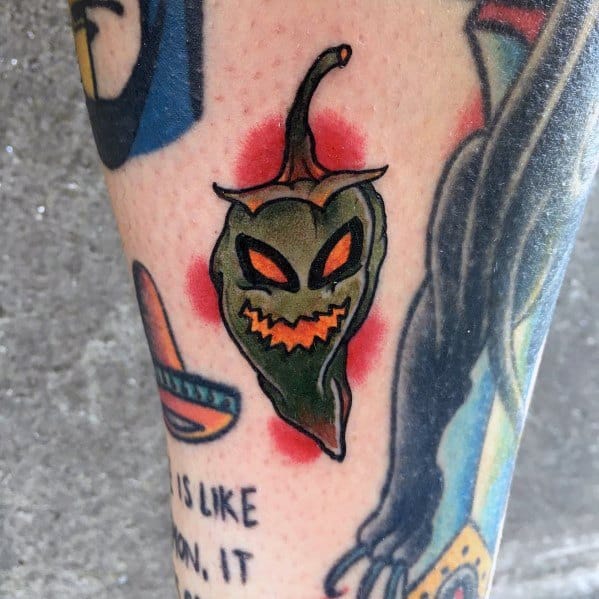 Green chili pepper tattoo with a sinister, jack-o'-lantern-style face, glowing eyes, and fiery red shading for a bold and spooky look.