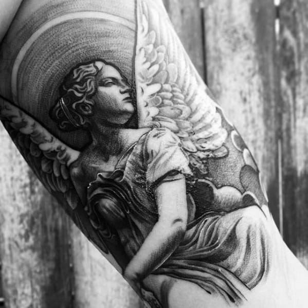 A black and grey tattoo on the forearm featuring an angel with large wings and a serene expression, with intricate shading details.