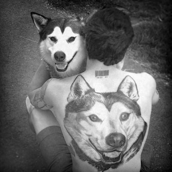Black and white photo of a man with a large realistic Siberian Husky tattoo on his back, perfectly aligned with his dog's face for a creative effect.