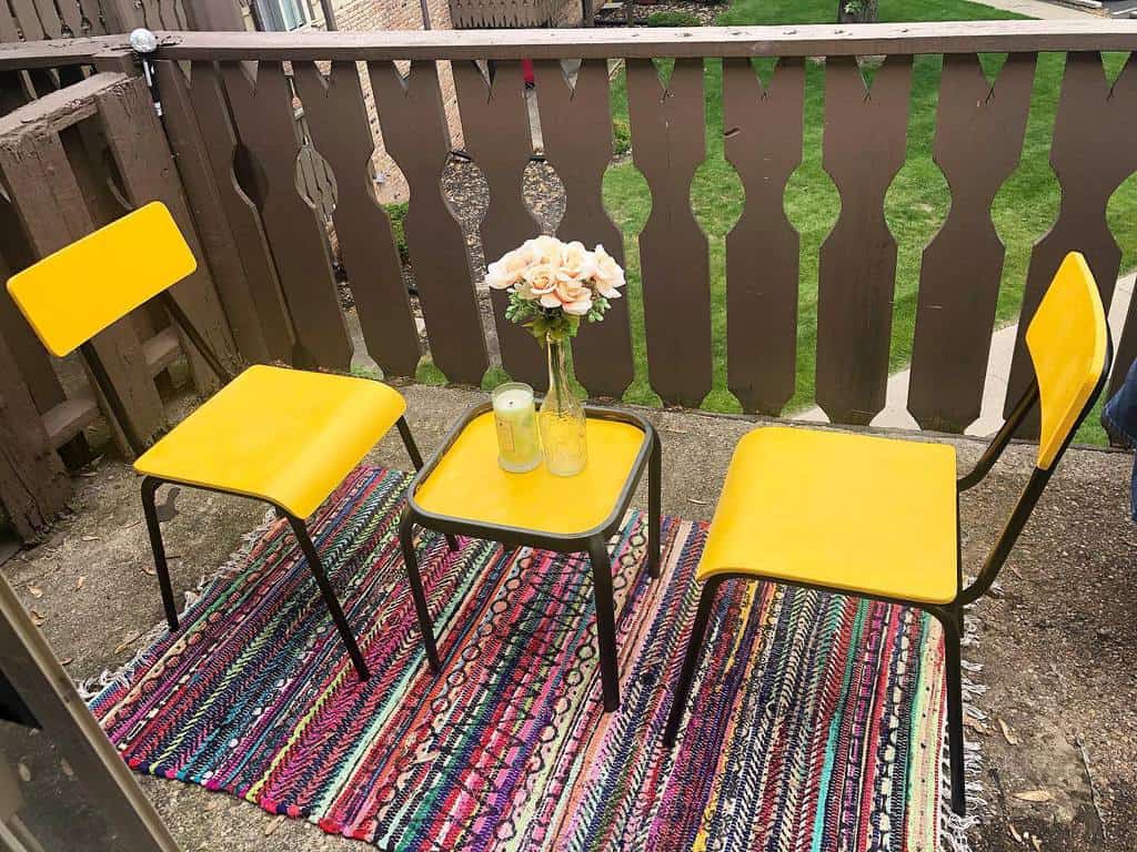 Small balcony with yellow chairs, matching table, colorful rug, and simple floral decor.