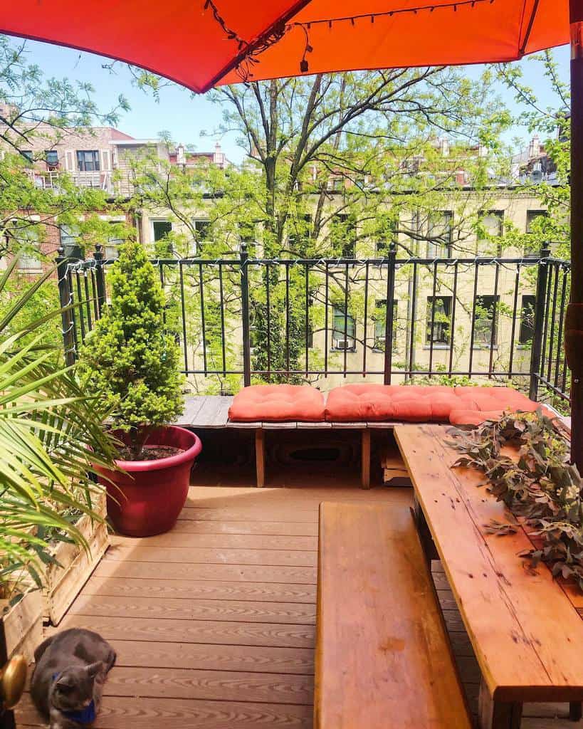 Quaint balcony with a wooden bench, table, potted plants, and a bright red umbrella for shade.