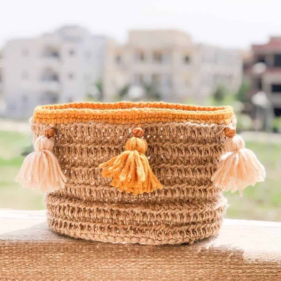 Woven basket with orange crochet trim and tassel accents displayed outdoors.