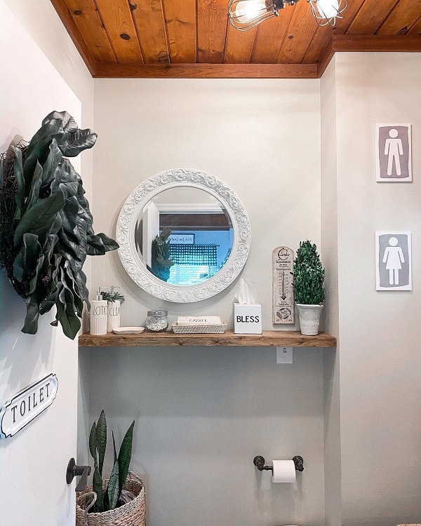 Cozy farmhouse bathroom with a circular mirror, wooden shelf, plants, and decorative signs against a white wall