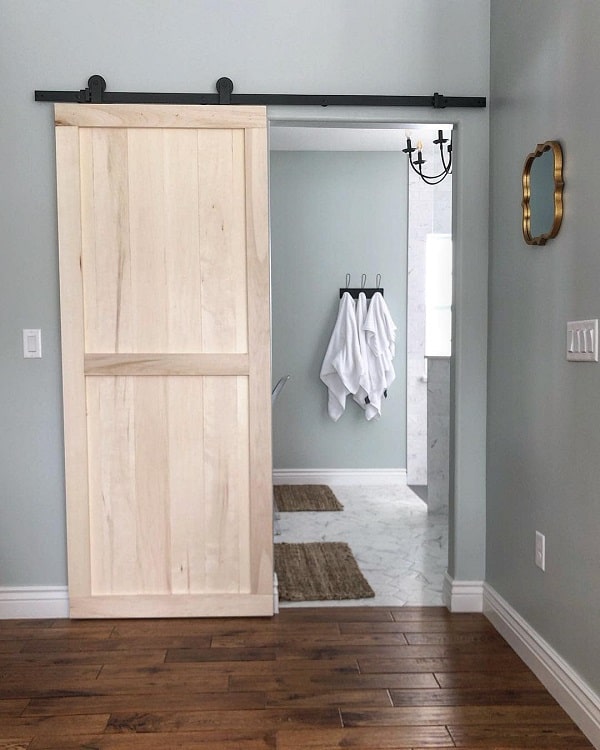 Light wood sliding barn door leading to a master bathroom with marble flooring and white towels hanging.