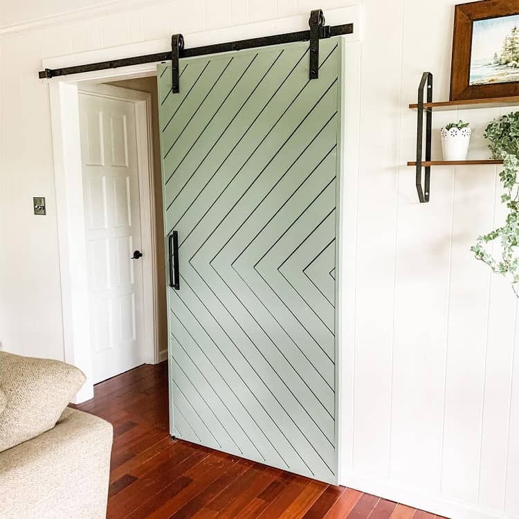 Light green sliding barn door with chevron pattern in a living room with wood floors and white walls.