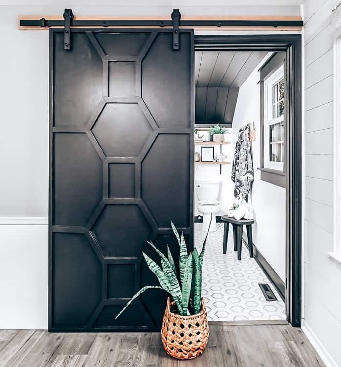 Black wooden sliding barn door with geometric panel design leading to a bathroom with hexagon tile flooring.