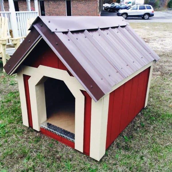 Dog house with a brown metal roof and red wooden walls, featuring a beige trim and a wide entrance, set in a grassy outdoor area