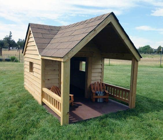 A wooden dog house with a porch and shingle roof, featuring two small chairs on the porch