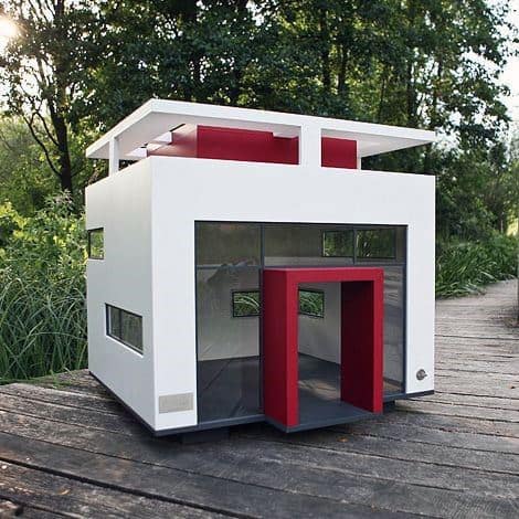 Modern, minimalist dog house featuring red and white accents, set on a wooden deck surrounded by lush greenery