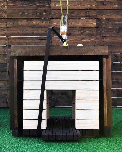 Modern dog house made from white-painted wooden slats, with a sleek design and a decorative vase on top, set on a green grass surface