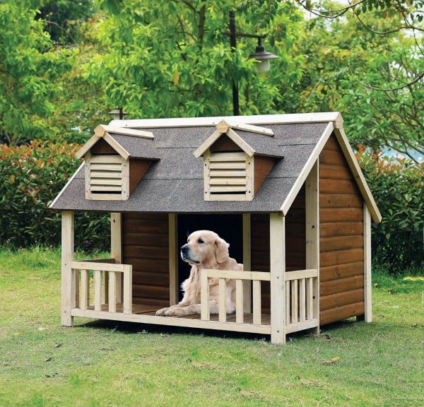 Dog lounging in a spacious wooden dog house with a front porch and two small windows, set in a lush green outdoor space
