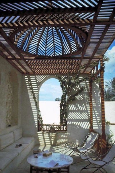 Ornate pergola with domed wood slats, arched wall, cozy seating, and intricate shadow patterns.